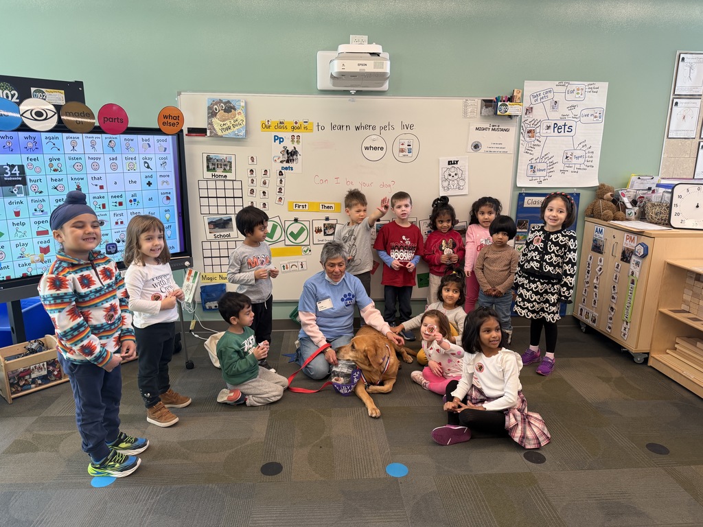 A class photo of students standing around the dog who is sitting on the floor with their owner.