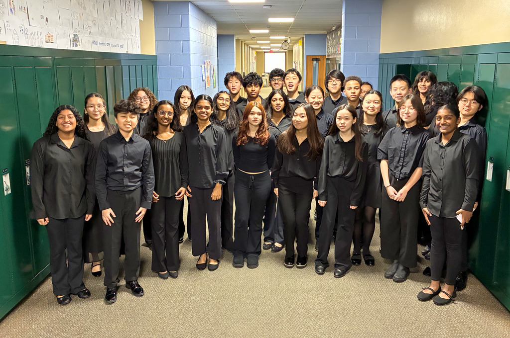 A group of chamber singers, standing in a group in a hallway all wearing black and smiling for a photo.