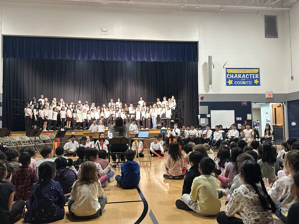 Students on stage, while others are sitting in chairs playing an instrument. STudents are sitting on the floor watching. 