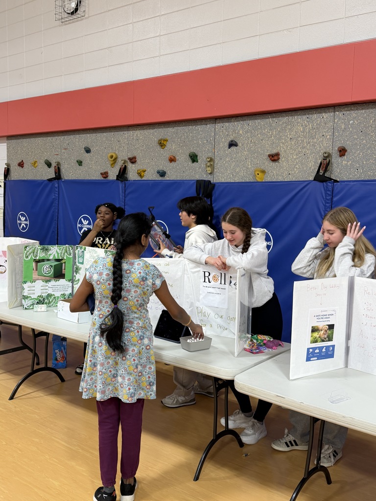 STudents standing behind tables with projects on them while another girl looks at their work.