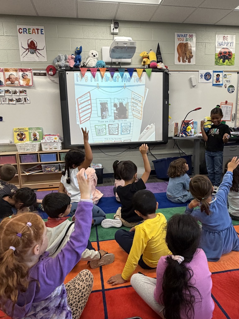 Student standing in front of the class pointing to students with their hands up.