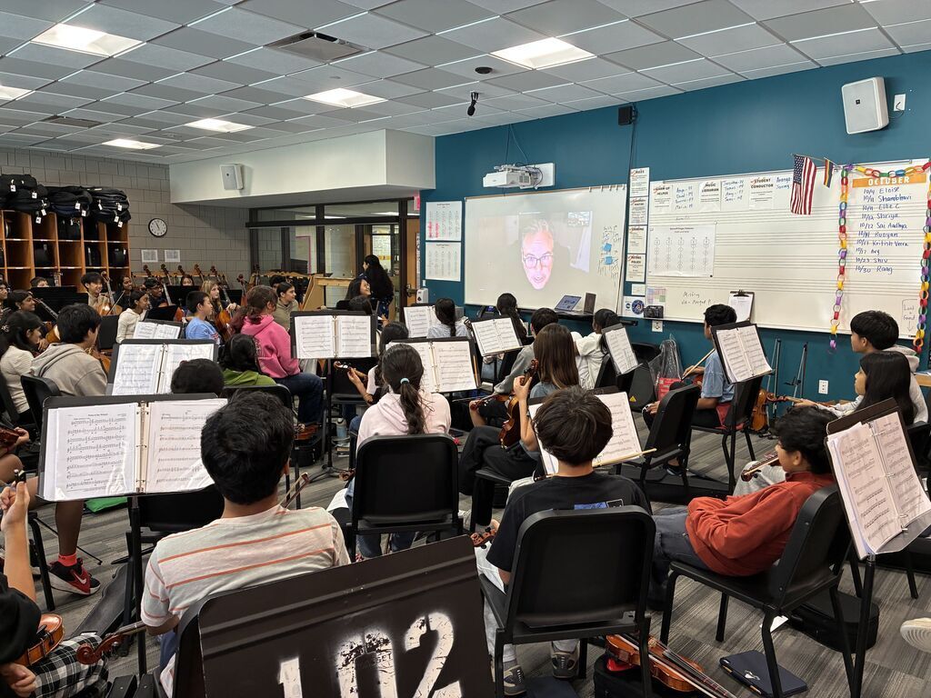 Students sitting in front of their instruments with a person on zoom presented on the board.
