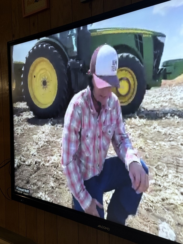 A tv screen showing an image of a farmer out in a field kneeling in front of a green tractor