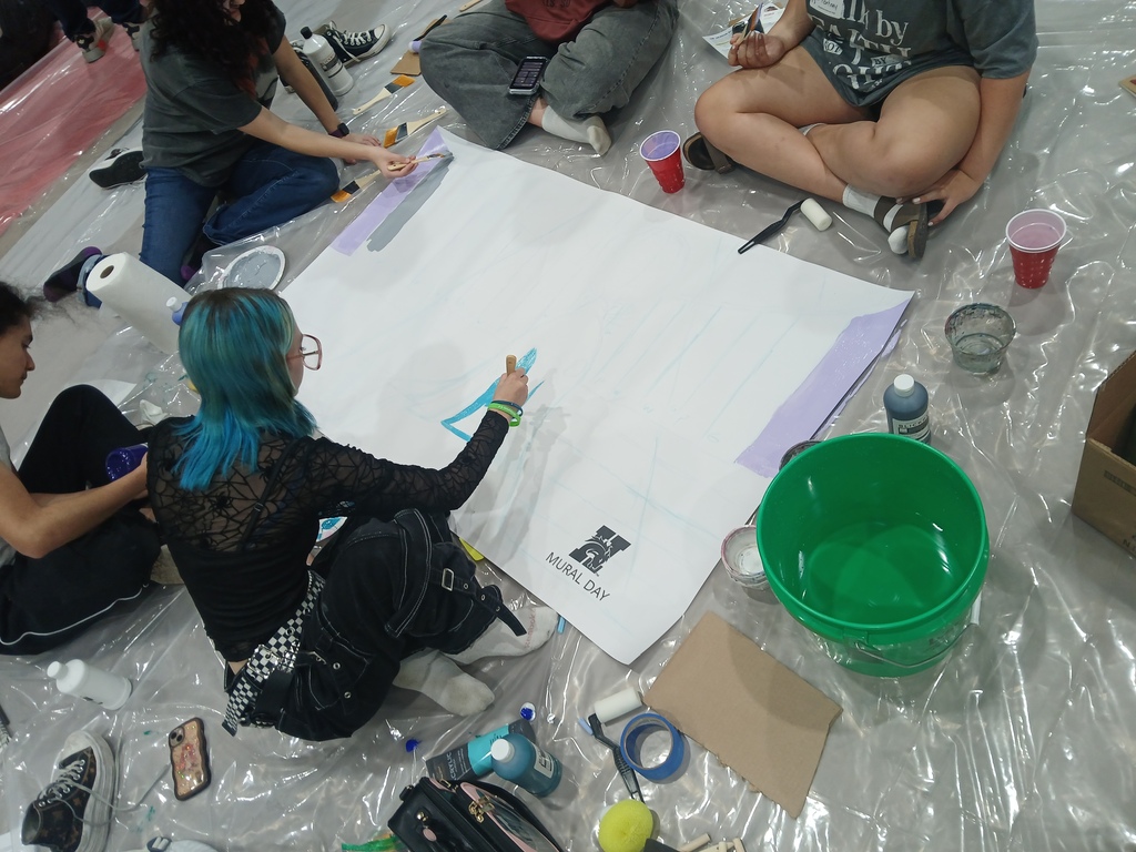 A picture of the students starting to add colors. So far, they've added some light purple and gray stripes, and a triangle of teal in the middle of the mural.