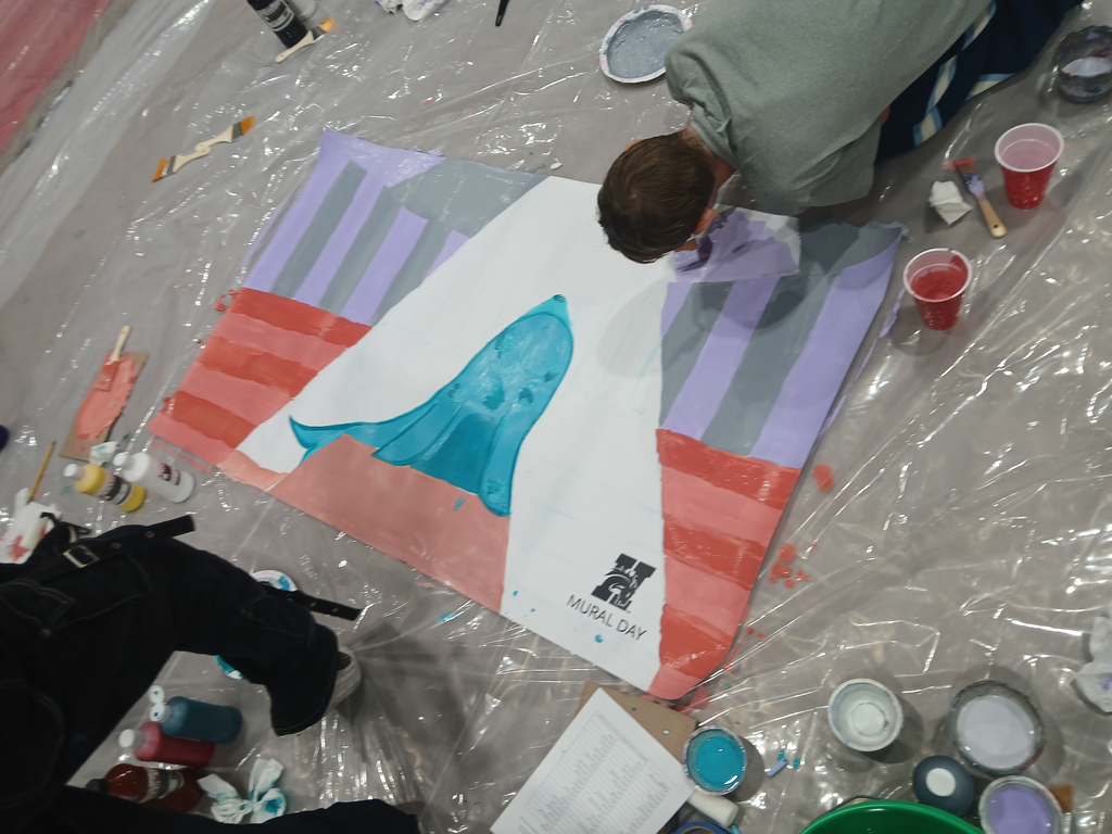 A picture of the mural halfway completed. There are light purple and grey circus tent strips in the background, red and pink stripes to make bleachers, and a teal seal in the middle of the page sitting on a pink podium. A cone of white sits around the seal where light will be painted later.