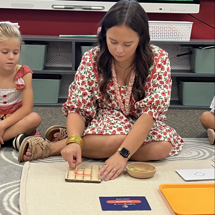 this photo is of an educator modeling to her students how to use tools for learning. The teacher is sitting on the floor, putting rubber bands on a board in shapes.