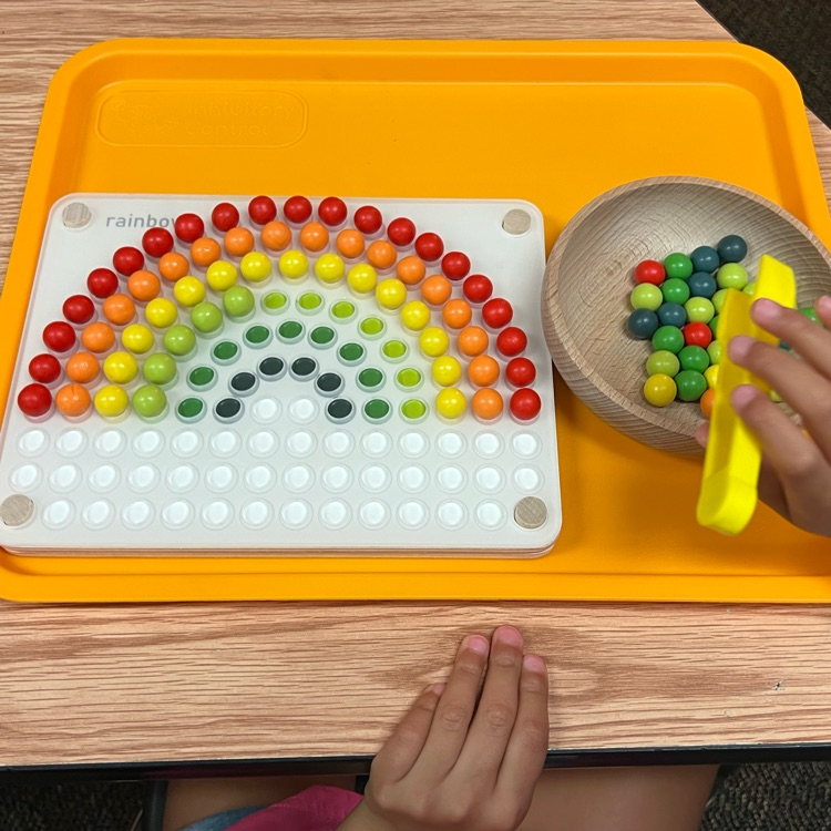 this picture is of the work students are doing at tables. The student is placing colored wooden balls in a rainbow pattern.