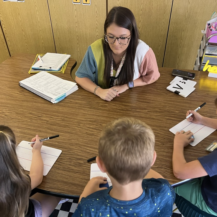 this teacher is sitting at a small group table with three young students. Each student has a whiteboard, a marker and eraser. They are working in a small group.
