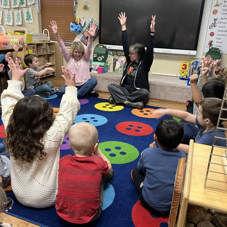 professional educator in front of class, demonstrating reaching to the sun. Children facing educator. Children are also reaching to the sun.