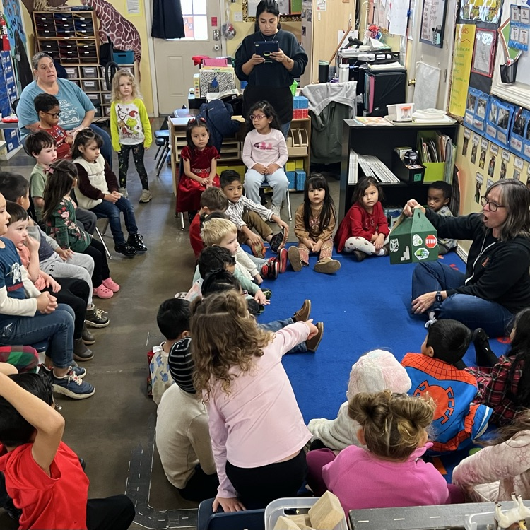 professional educator sitting on the carpet in front of a group of children. Educator is holding up a box that contains props for a story.