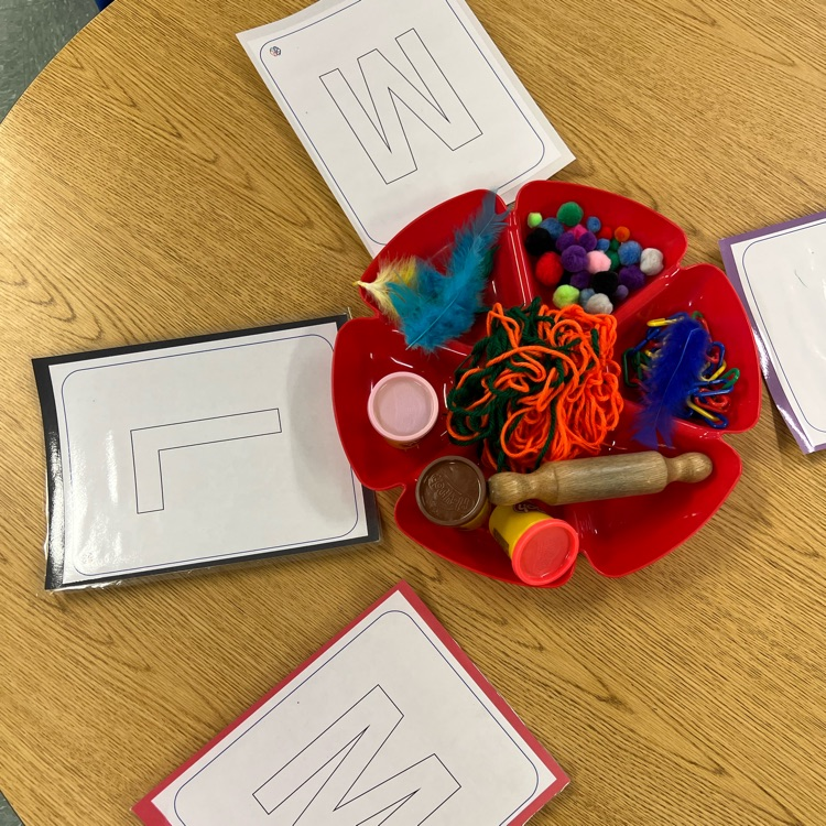 Tabletop literacy activity with uppercase letter cards and a tray of fine motor materials including pom-poms, yarn, feathers, and play dough tools.