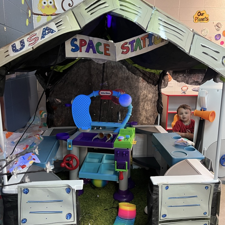 Pretend play “USA Space Station” center in a preschool classroom with a toy control panel, telescope, and space-themed decorations for dramatic play.