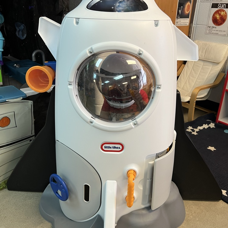 Child standing inside a white toy rocket in a preschool classroom dramatic play area designed for a space exploration theme.