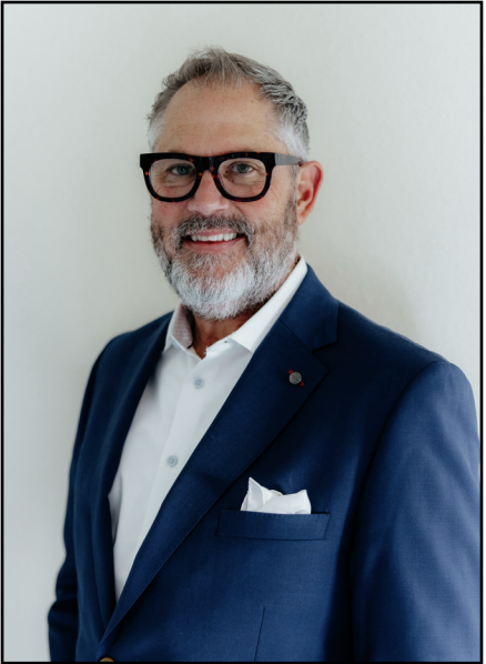A smiling man with glasses and a beard is wearing a blue suit and white shirt. He stands against a plain light background, exuding a professional demeanor.