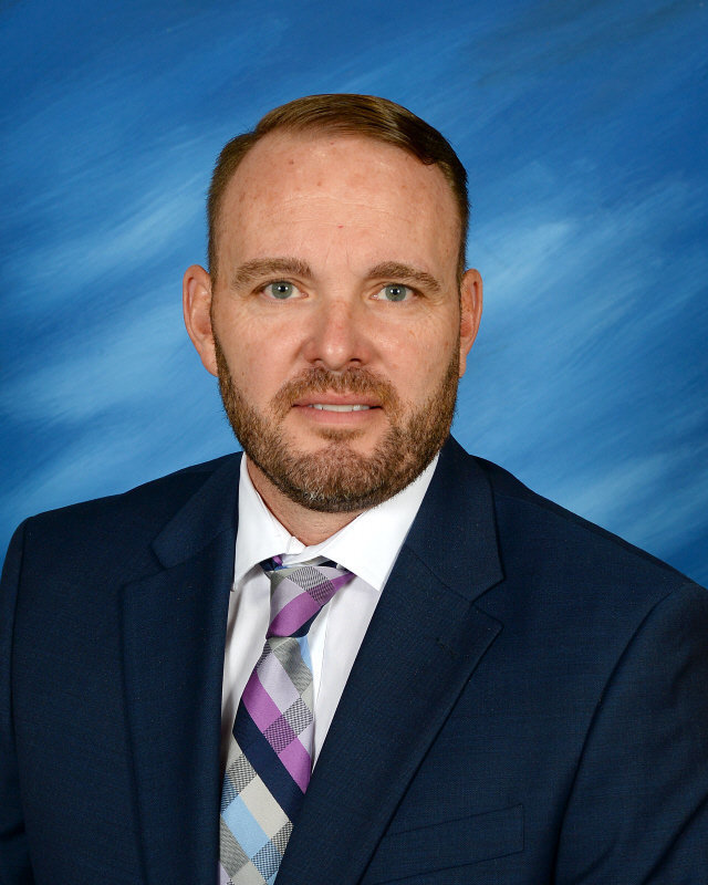 Man with short hair and a beard in a blue suit and multicolored plaid tie, posing against a blue background. He has a serious expression.