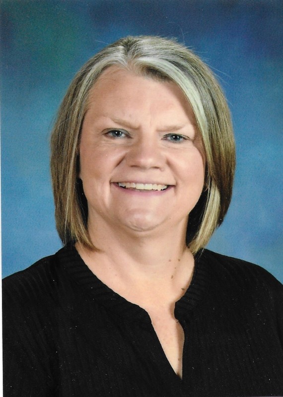 Smiling woman with short blond hair wearing a black top against a blue background. She exudes a friendly and approachable demeanor.