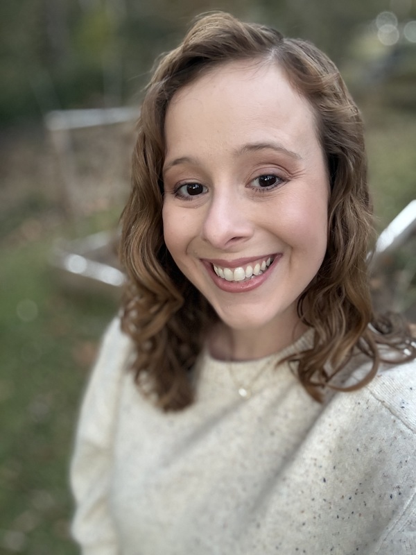 Smiling woman with curly brown hair wearing a light sweater, outdoors. The background is blurred greenery, creating a warm and cheerful atmosphere.