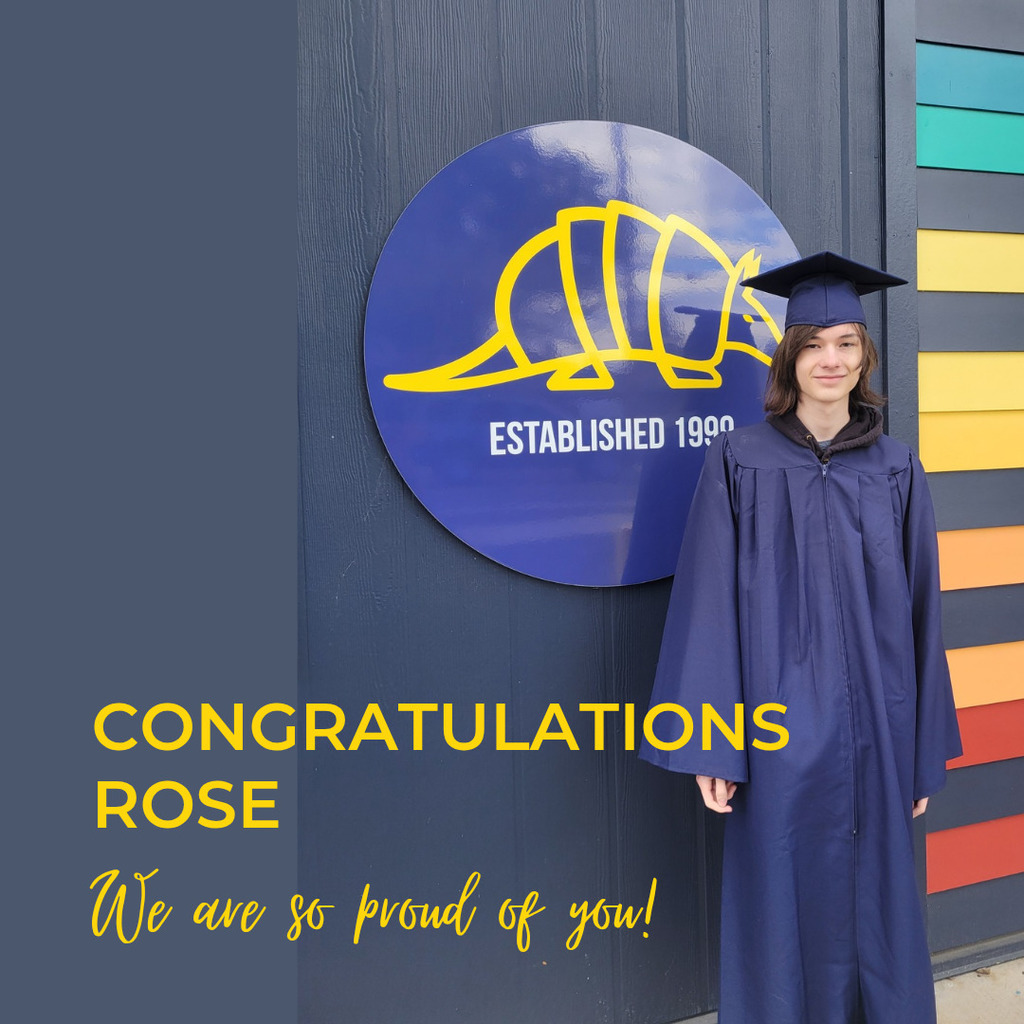 student in cap and gown in front of school sign with yellow wording on blue background