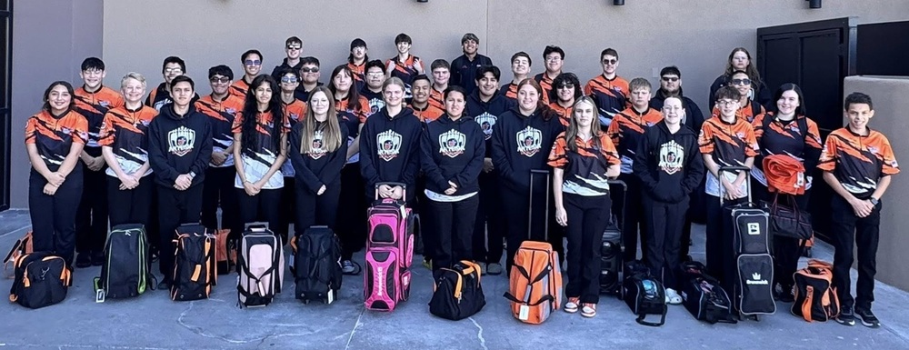 A large group of male and female teens pose for a photo outside a bowling center with a row of bowling bags lined up in front of them.