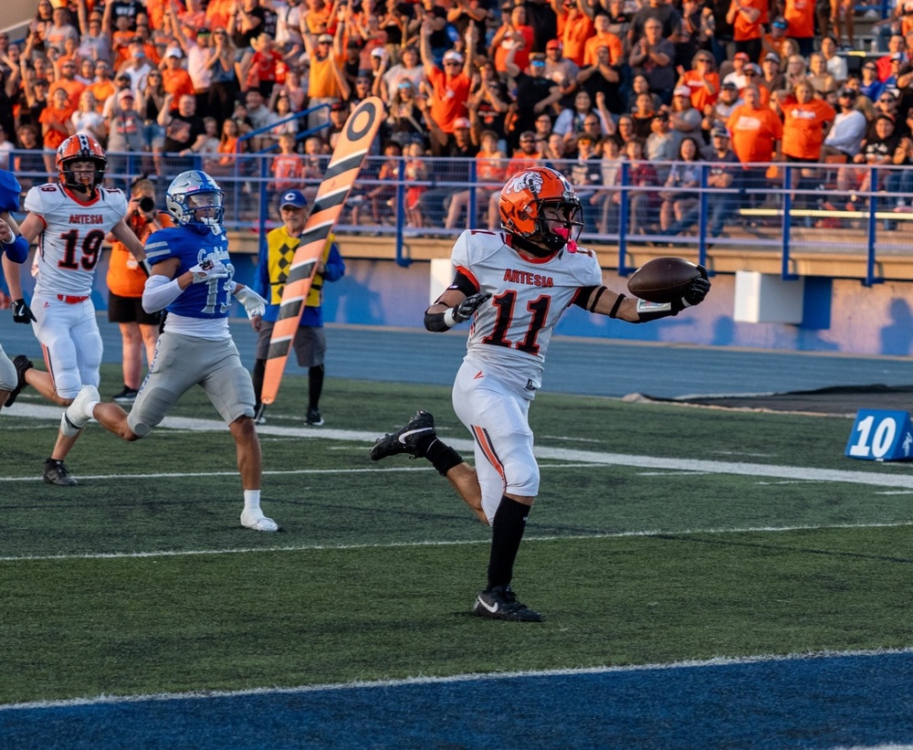 A teen boy in a white-and-orange football uniform holds the ball in front of him in his left hand as he runs over the goal line. Behind him, a cheering crowd dressed in orange can be seen.