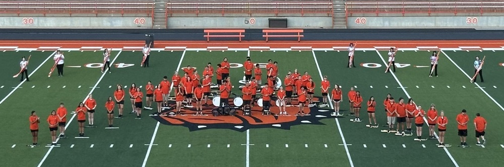 A high school marching band clad in orange t-shirts and black shorts stand in an A formation on a football field.