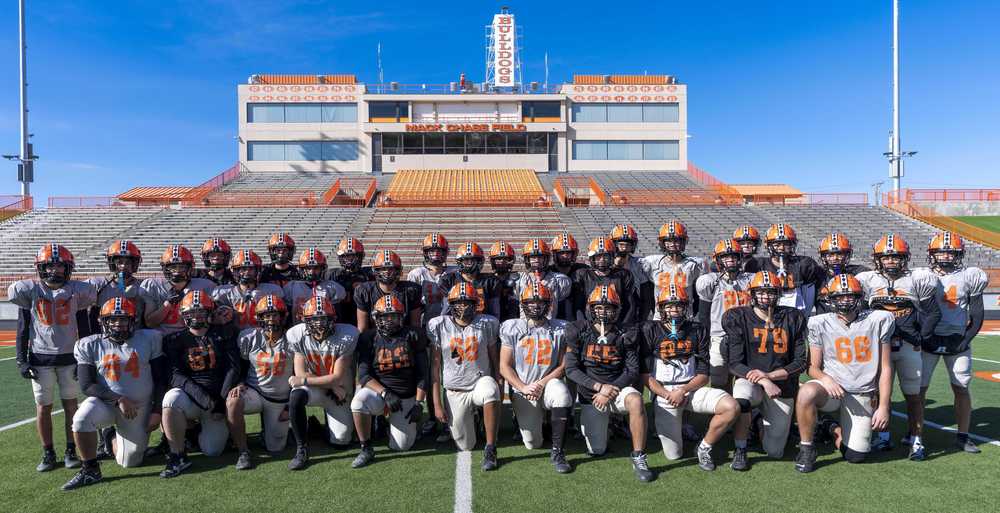 A large group of teen boys in football helmets, some in black jerseys and some in grey jerseys, pose for a photo on a football field.