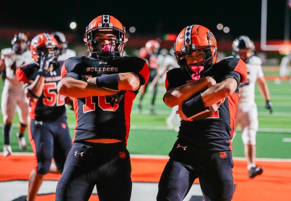 Two teen boys in black-and-orange football uniforms pose for a photo with their arms folded across their chests. One holds a football in his arms.