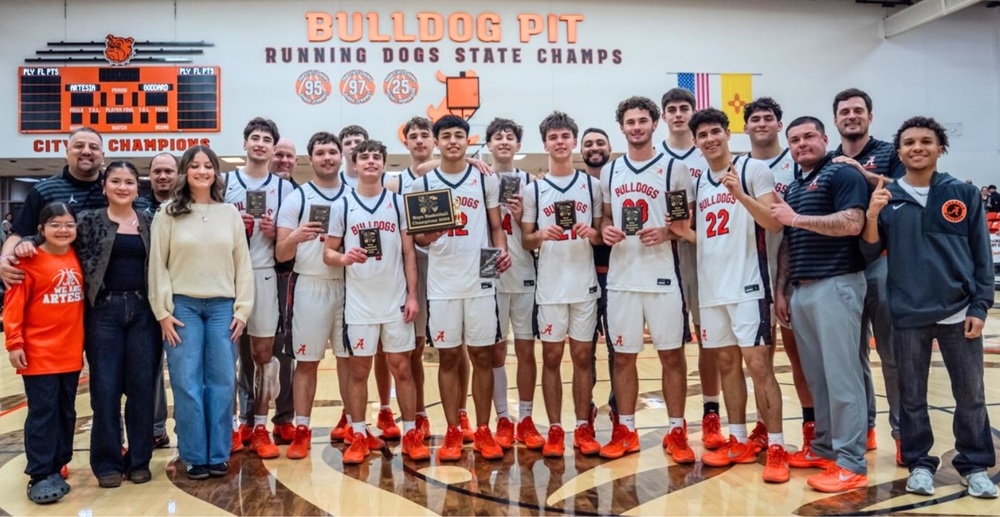 A boys' basketball team in white and orange uniforms with orange shoes poses for a photo holding a trophy and individual plaques, flanked by coaches and managers.
