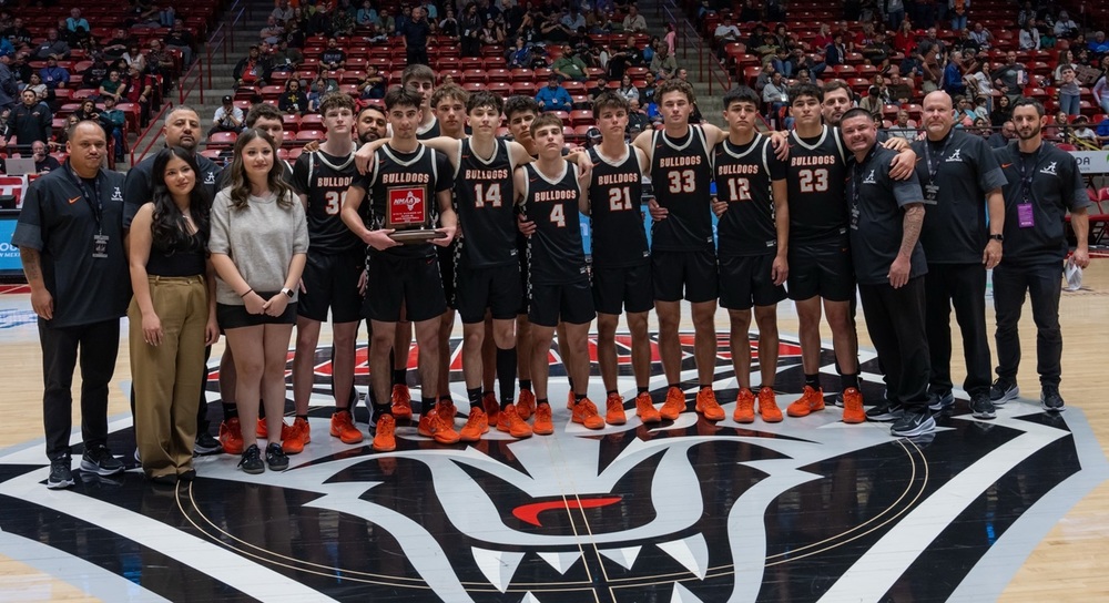 A boys' basketball team in black and orange uniforms poses for a photo with their coaches. One player holds a red trophy.