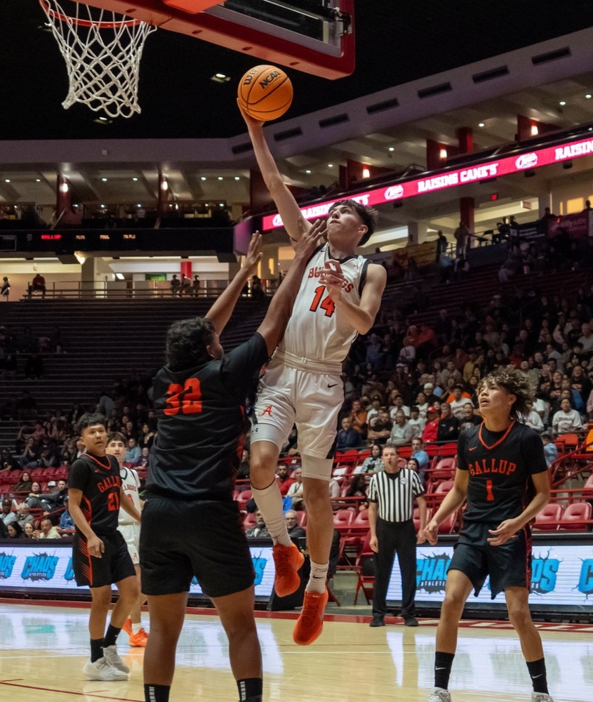 A teen boy in a white and orange basketball uniform goes high in for a layup as an opponent in black and orange raises both arms in front of him. 