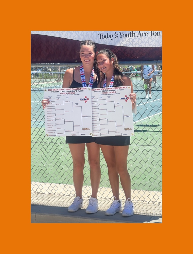 Two teen girl pose for a photo in front of tennis courts, holding large brackets and wearig medals around their necks.