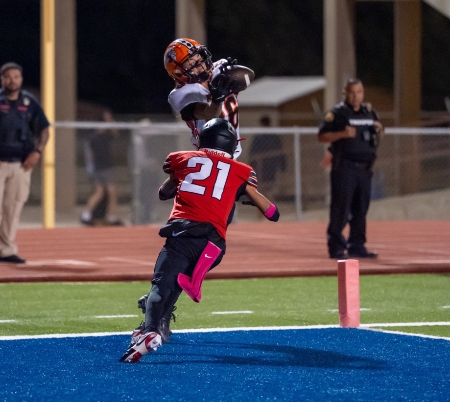 A teen boy in a white-and-orange football uniform leaps into the air to catch a football over top of an opponent in red and black.