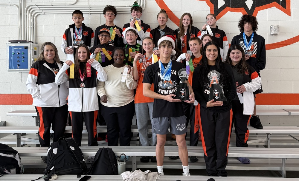 A large group of male and female teens pose in a set of bleachers with their female coach. Several students hold up ribbons and one boy and one girl in front display trophies.