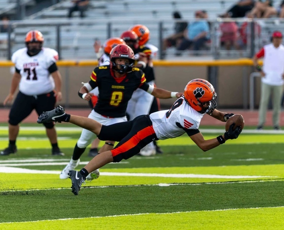 A teen boy in an orange, white and black football uniform dives to catch the football in both hands as an opponent in black, red and gold looks on.