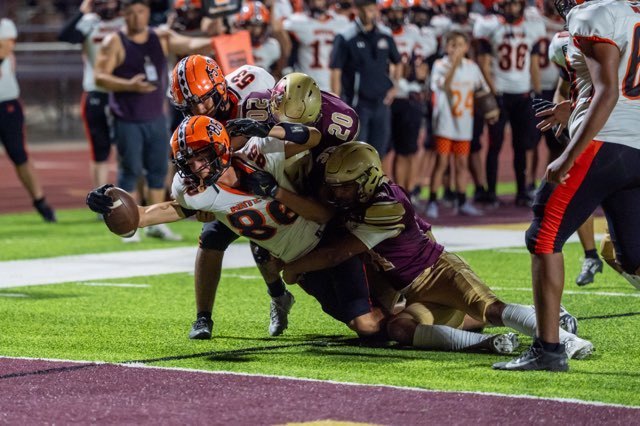 A teen boy in a white, orange and black football uniform extends the ball over the goal line with his right hand as three defenders hang from his body.