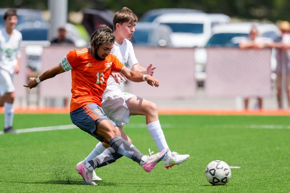 A teen boy in an orange-and-grey soccer uniform kicks the ball past an opponent in all white.
