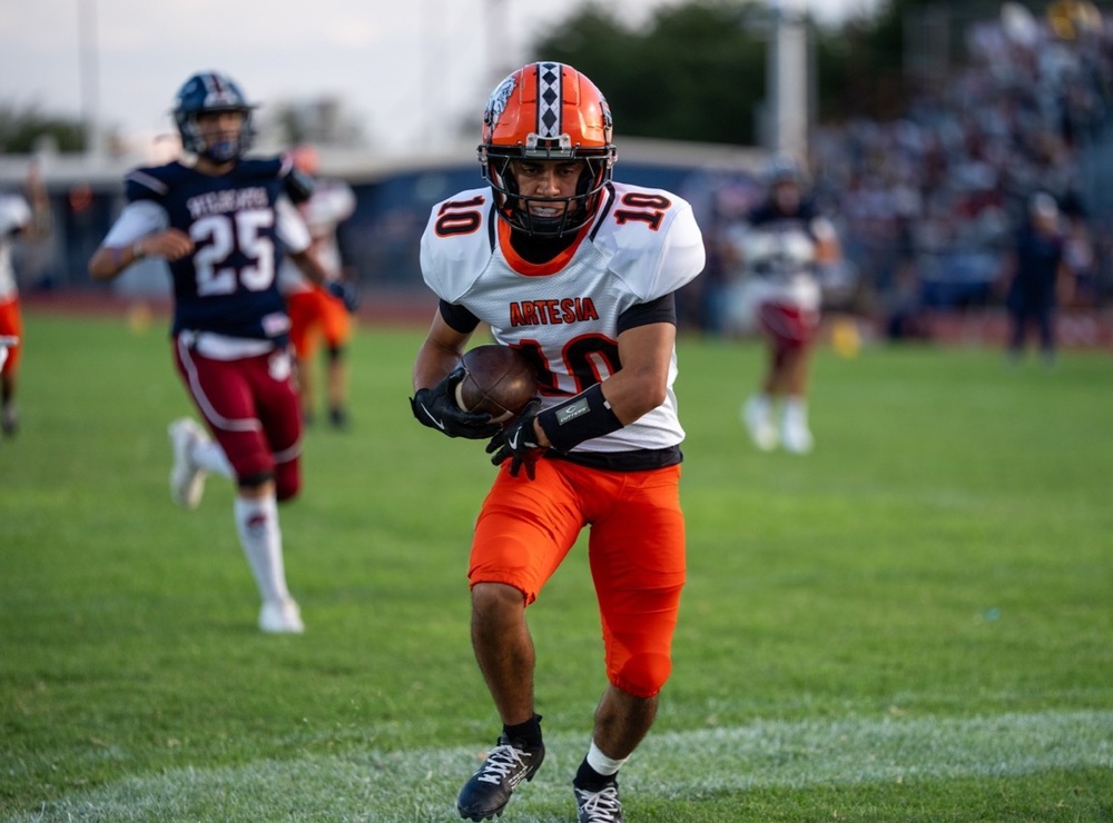 A teen boy in a white-and-orange football uniform cradles the ball in his right arm and looks straight ahead as he turns into the end zone.