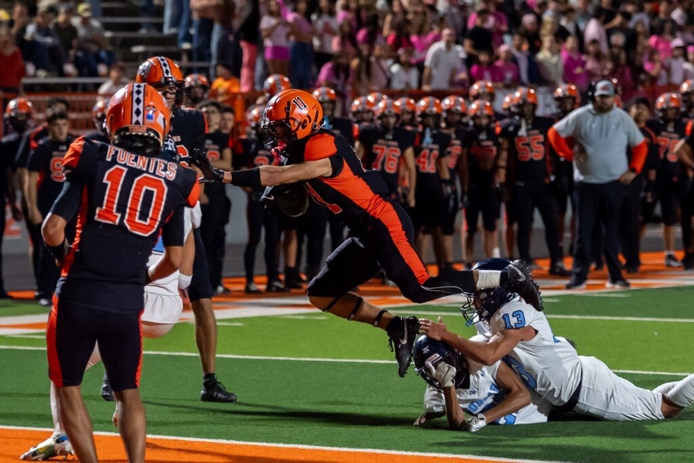 A teen boy in a black-and-orange football uniform goes airborne toward the goal line to avoid a pair of diving opponents in white and baby blue.