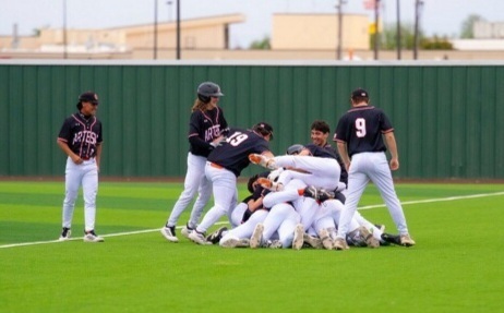 A boys' baseball team in black, orange and white uniforms dog piles in right field after a win.