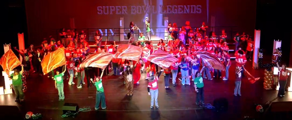 A group of male and female teens in football jersey t-shirts, some waving orange flags, dance and sing on a stage.