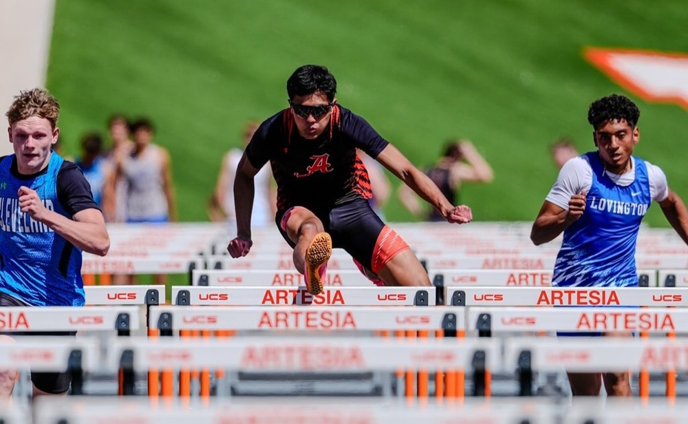 A teen boy in a black and orange track uniform and sunshades clears a hurdle as two opponents in blue run on either side of him.