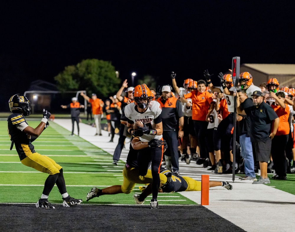 A teen boy in an orange, black and white football uniform runs across the goal line with the football. One defender in black and gold hangs from his waist and another is standing nearby, while the cheering sideline can be seen in the background.