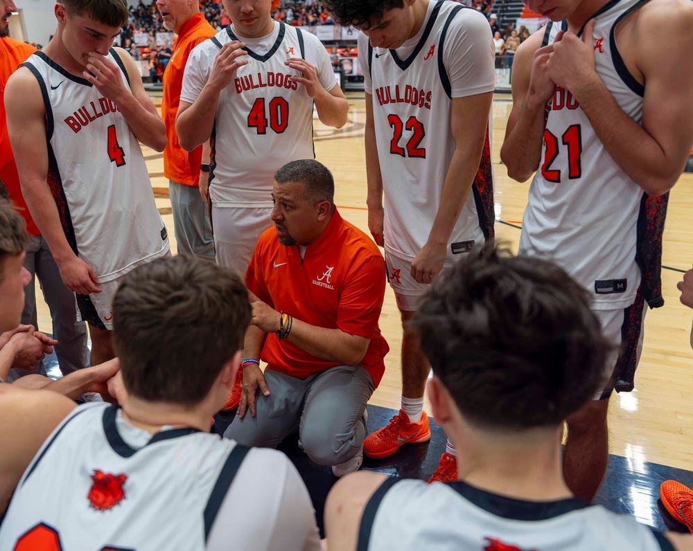 A group of teen boys in orange and white basketball uniforms gather around a male coach in an orange shirt as he speaks to them.