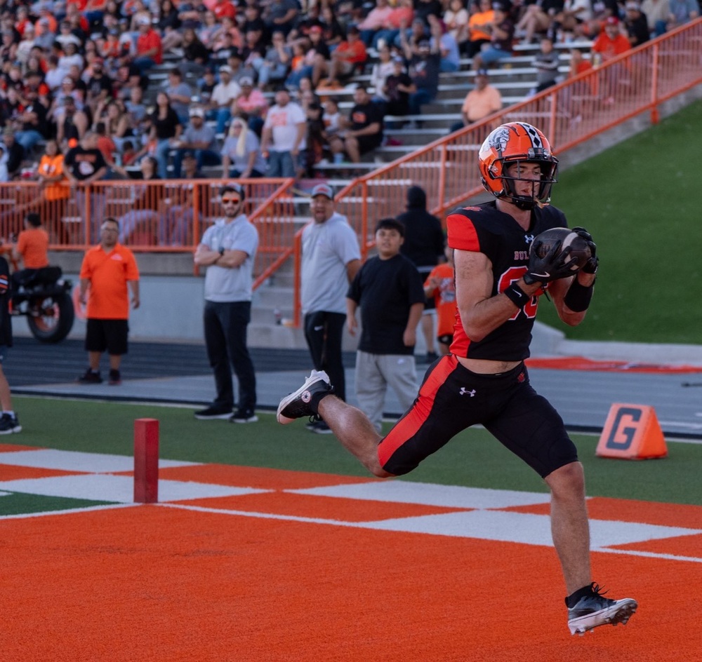 A teen boy in a black-and-orange football uniform runs through an orange end zone holding a football. A crowd seated in bleachers can be seen behind him.