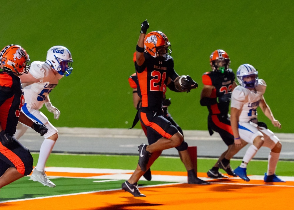 A teen boy in a black-and-orange football uniform runs into the end zone with the ball in his left hand as he raises his right fist in celebration.