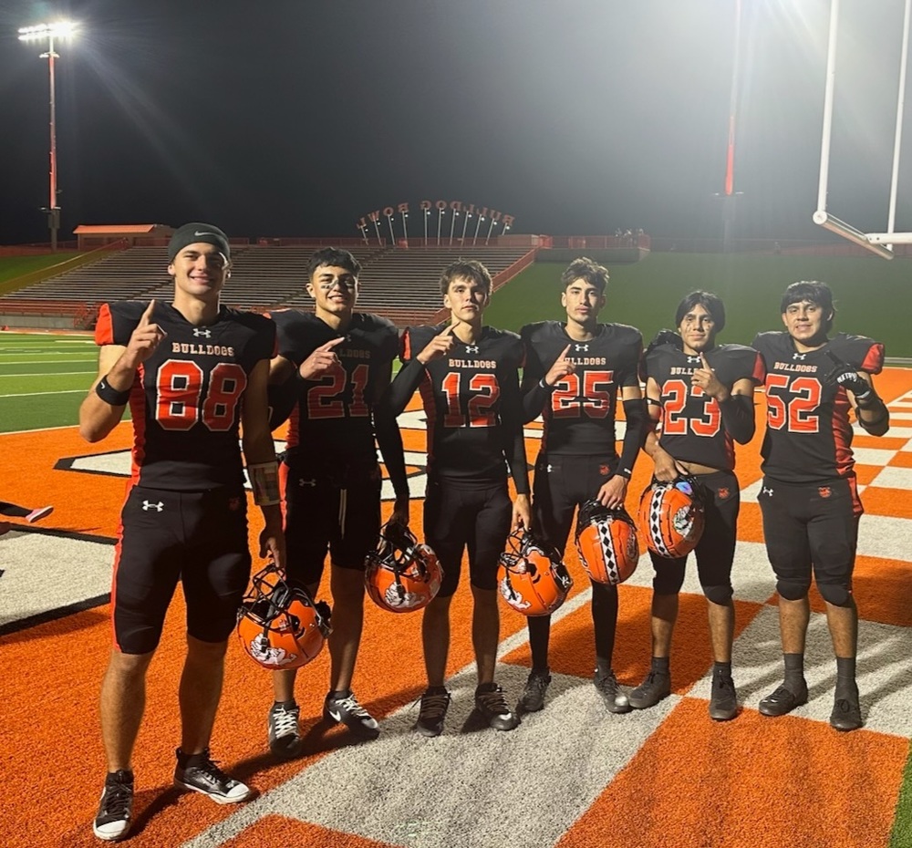 Six teen boys in black-and-orange football uniforms stand in a line in an orange-and-white checkerboard end zone and hold up number-one fingers.