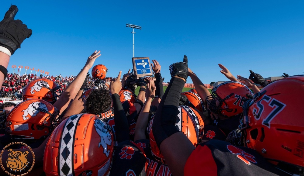 A large group of teen boys in orange football helmets hold their hands up around a blue trophy in the shape of the state of New Mexico.