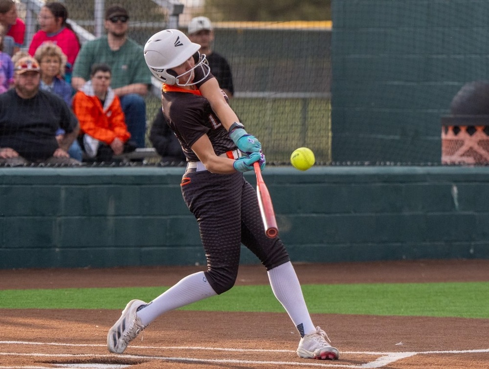 A teen girl in a black and orange softball uniform swings as she prepares to hit the ball.