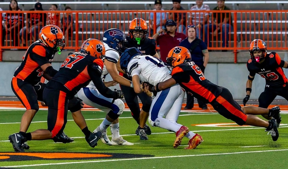 A teen boy in an orange and black football uniform dives to sack the opposing quarterback in white and blue as three teammates move in to assist.