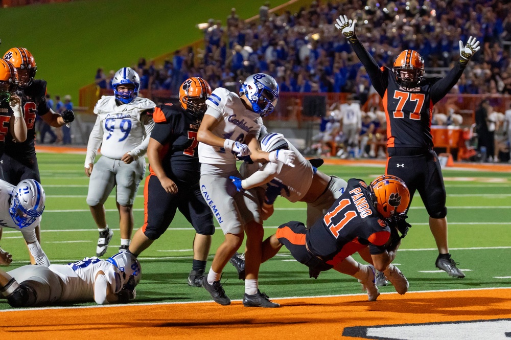 A teen boy in a black and orange football uniform dives into an orange end zone as opponents in silver and blue attempt to tackle him. A teammate standing near the goal line signals the touchdown.